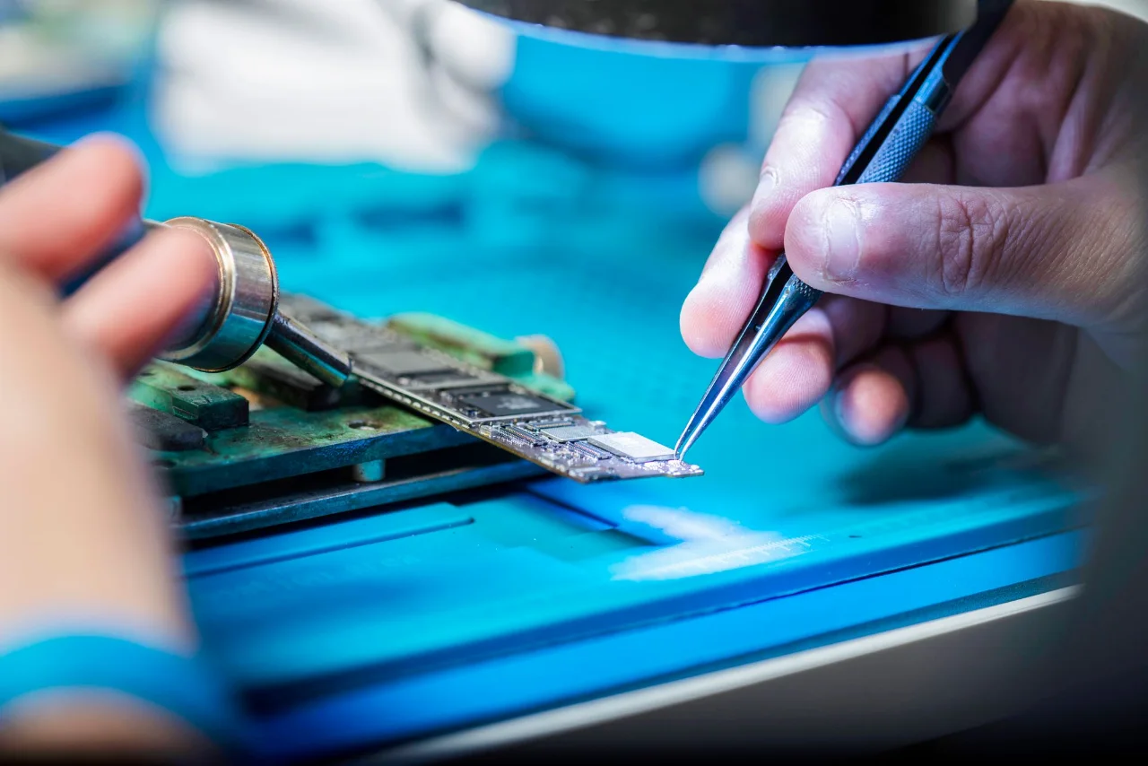 Hands using tools to repair a circuit board on a blue work mat